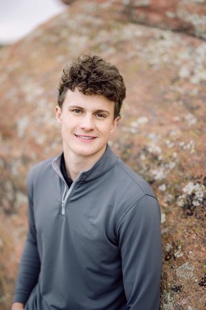Young man with curly hair smiling outdoors, wearing a gray quarter-zip pullover against a rocky background.