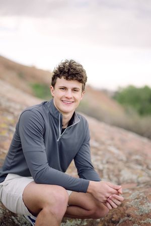 Smiling teenage boy in gray quarter-zip and shorts sitting on rocks outdoors with hills in background.