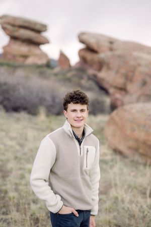 Young man in beige fleece jacket smiling outdoors at Red Rocks Park with iconic sandstone formations in background.