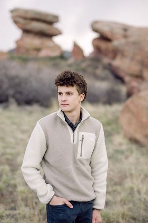 Young man in cream fleece pullover stands before red rock formations at Red Rocks Park, Colorado.
