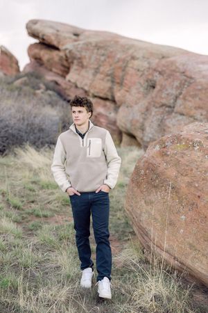 Young man in beige fleece and dark jeans stands confidently among red rock formations on an overcast day.