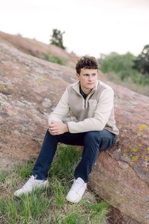 Young man in beige pullover and dark jeans sitting on a rock outdoors, posing for a senior portrait photo.