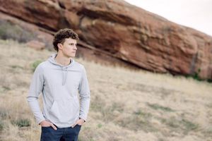 Young man in gray hoodie stands in front of red rock formations in a scenic outdoor landscape.