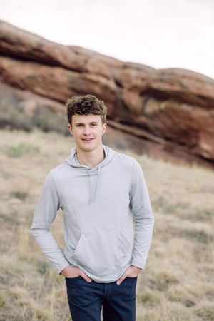 A young man in a gray hoodie and jeans stands smiling outdoors near red rock formations.