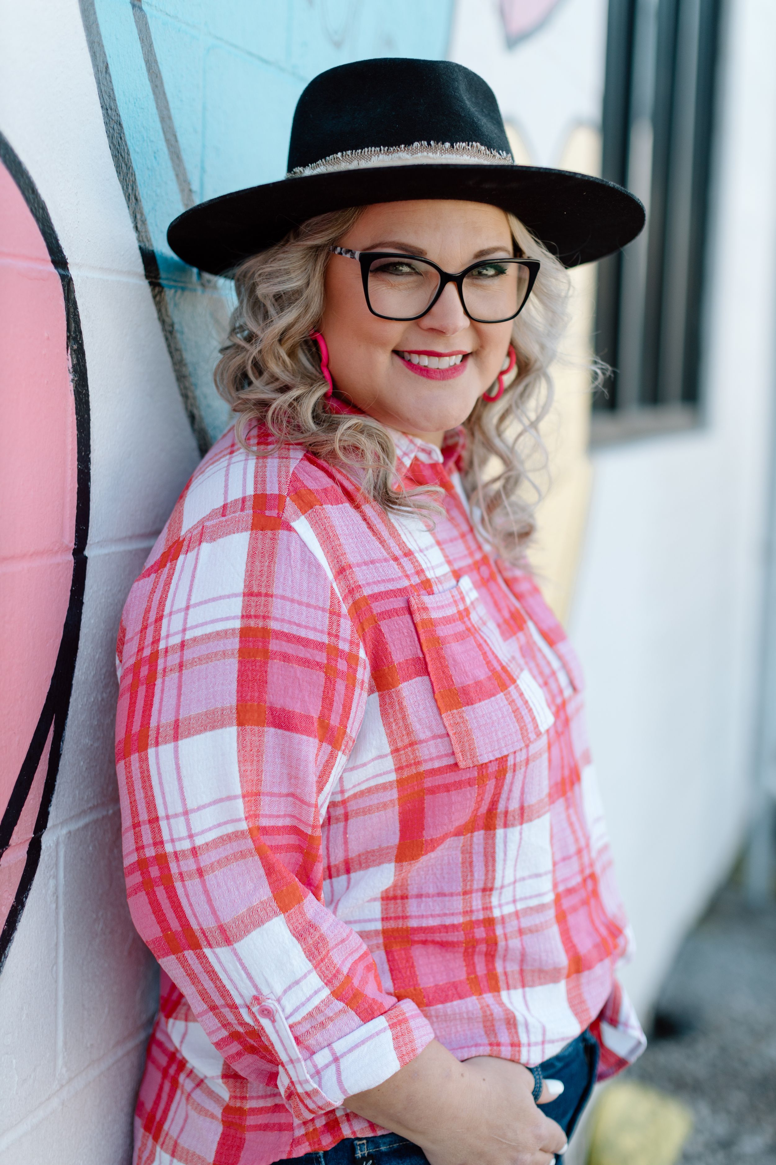A stylish portrait of someone in a pink plaid shirt, black hat with glasses leaning against a wall outdoors.