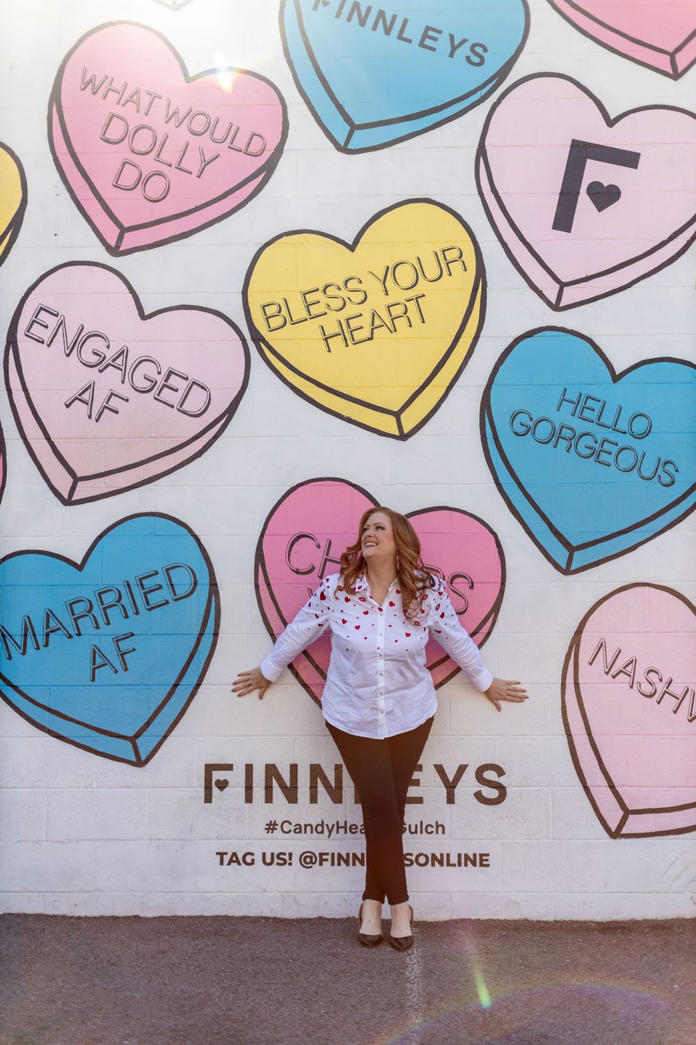 Someone poses in front of a colorful wall mural featuring candy heart designs with text at Finnleys retail store.