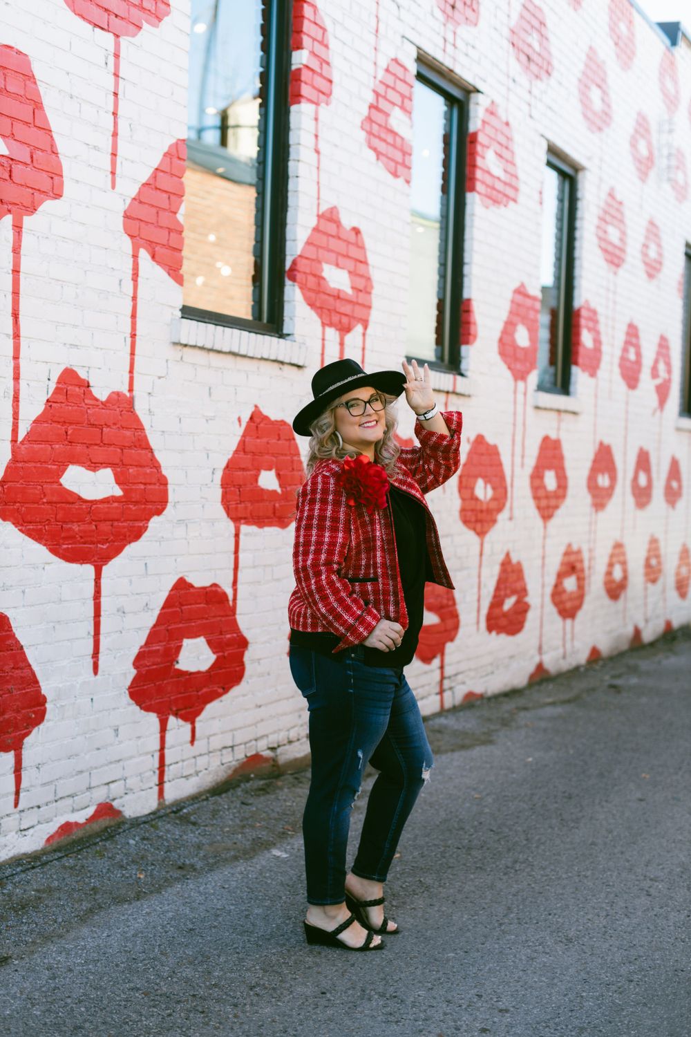 Artistic wall mural features repeating red lip prints on white brick wall with someone posing in plaid and a hat.