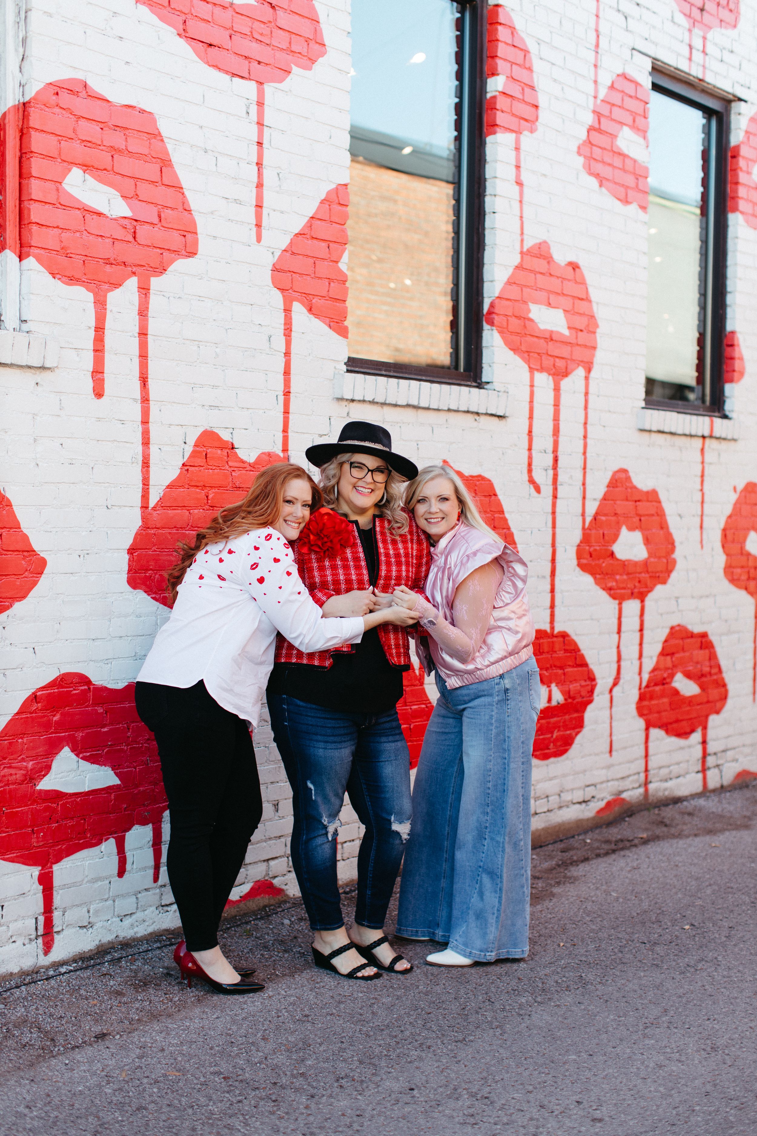 Three friends pose together against a white wall decorated with large painted red poppies in an urban setting.