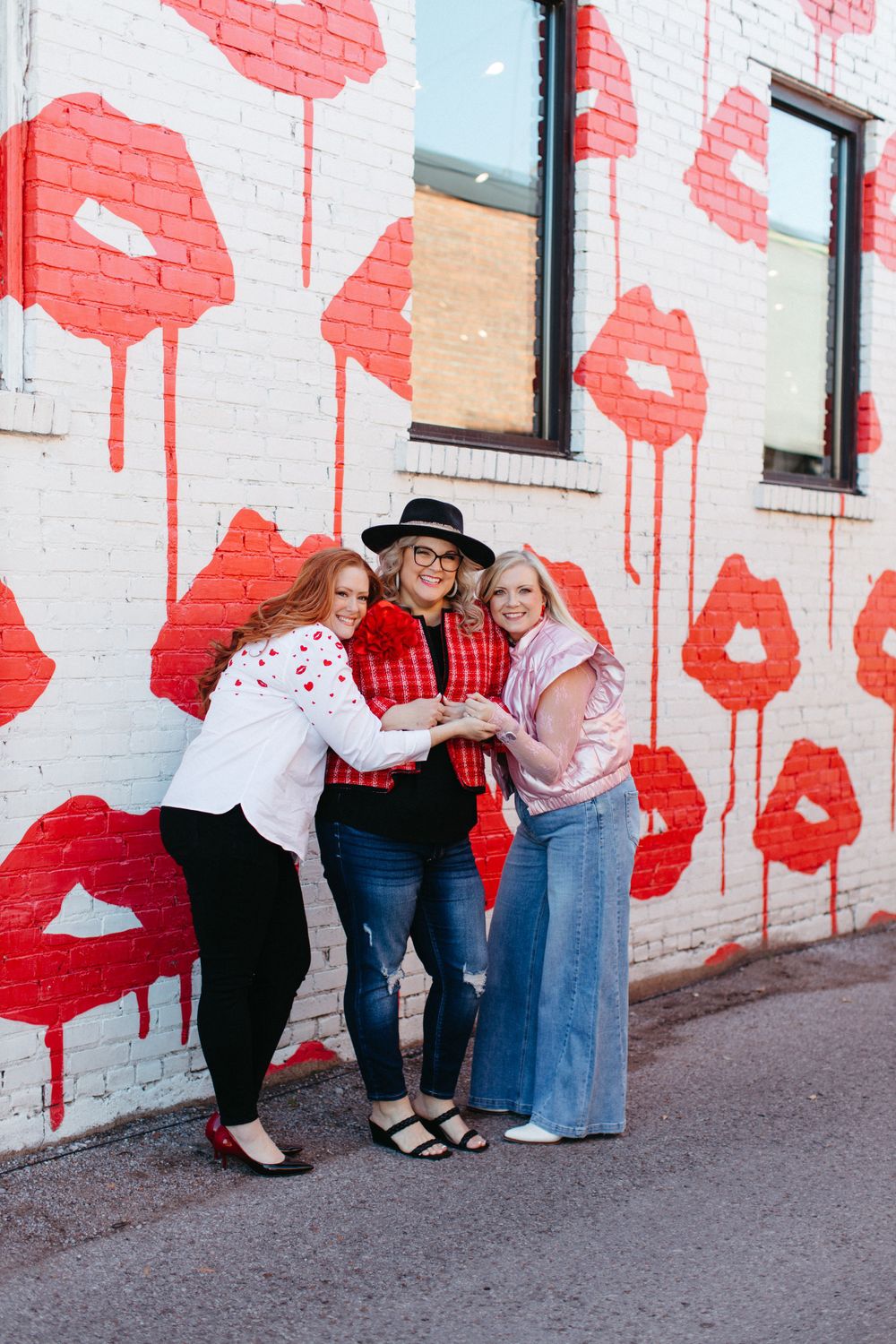 Three friends pose together against a white wall decorated with large painted red poppies in an urban setting.