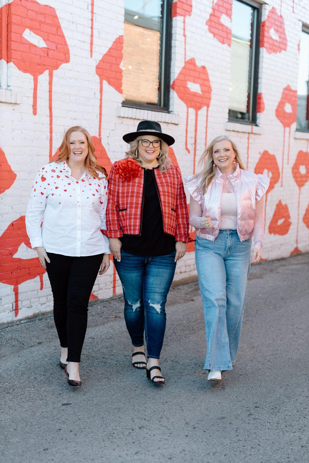 Three women walk side by side against a white wall decorated with red lipstick kiss patterns.