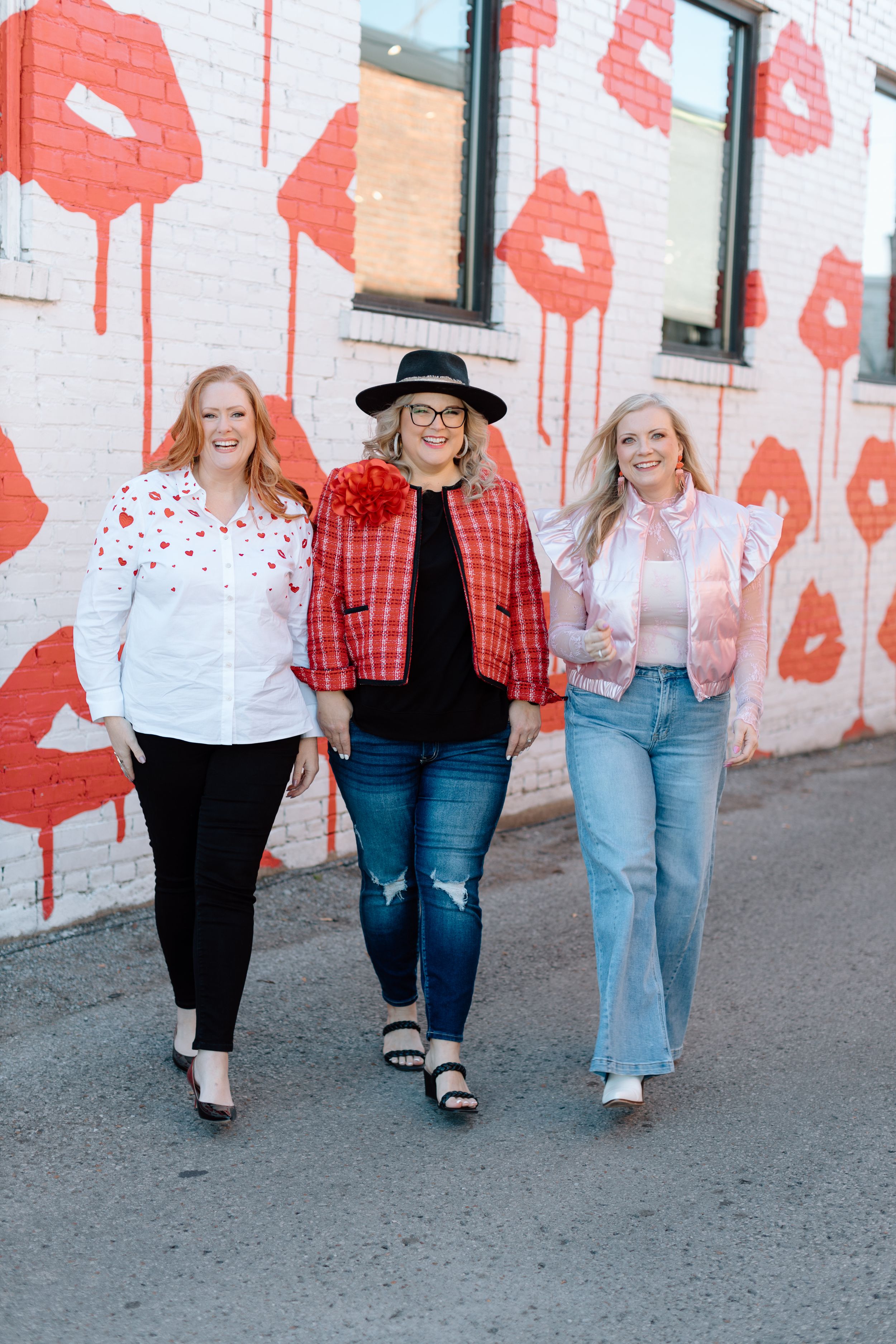 Three women walk side by side against a white wall decorated with red lipstick kiss patterns.