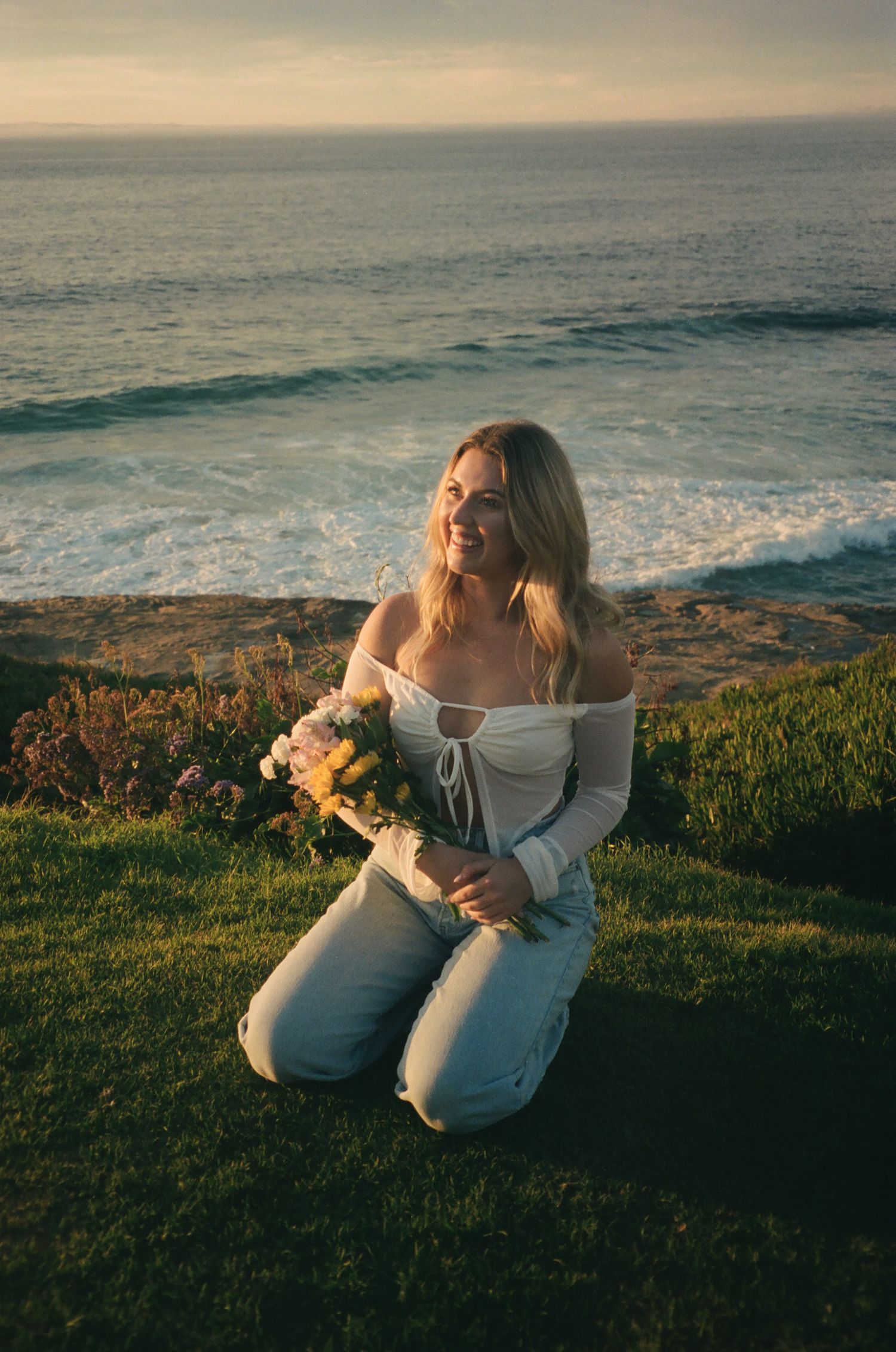 A blonde woman kneels on the grass overlooking the ocean holding a bouquet of flowers and wearing jeans and a white top