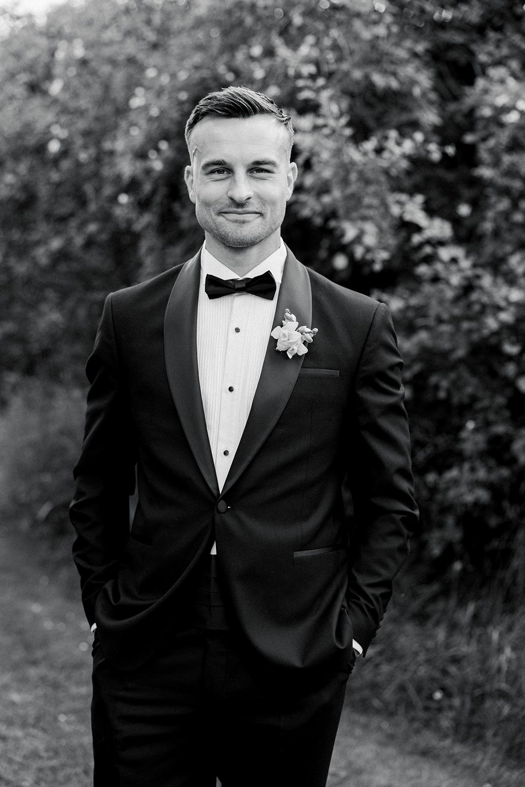 A groom in a classic black tuxedo and bowtie poses outdoors against a natural foliage background.