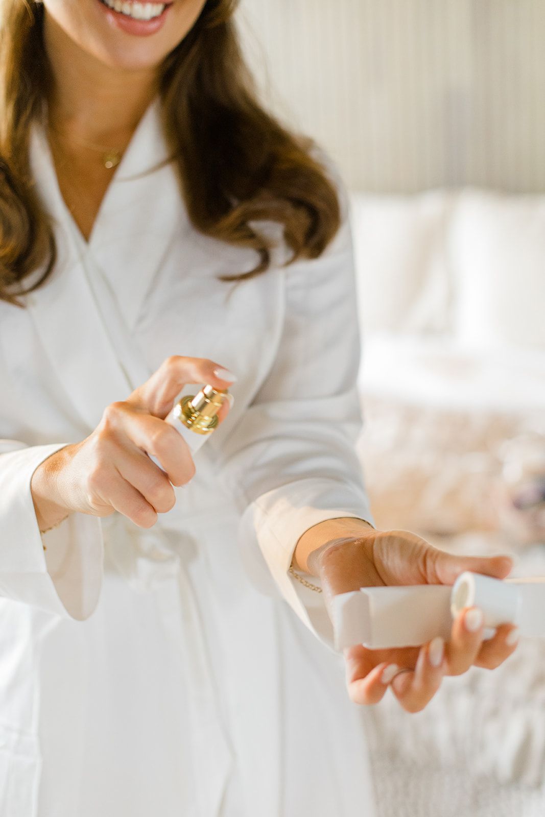 Woman in white robe spraying perfume in a bright, airy setting with natural light.