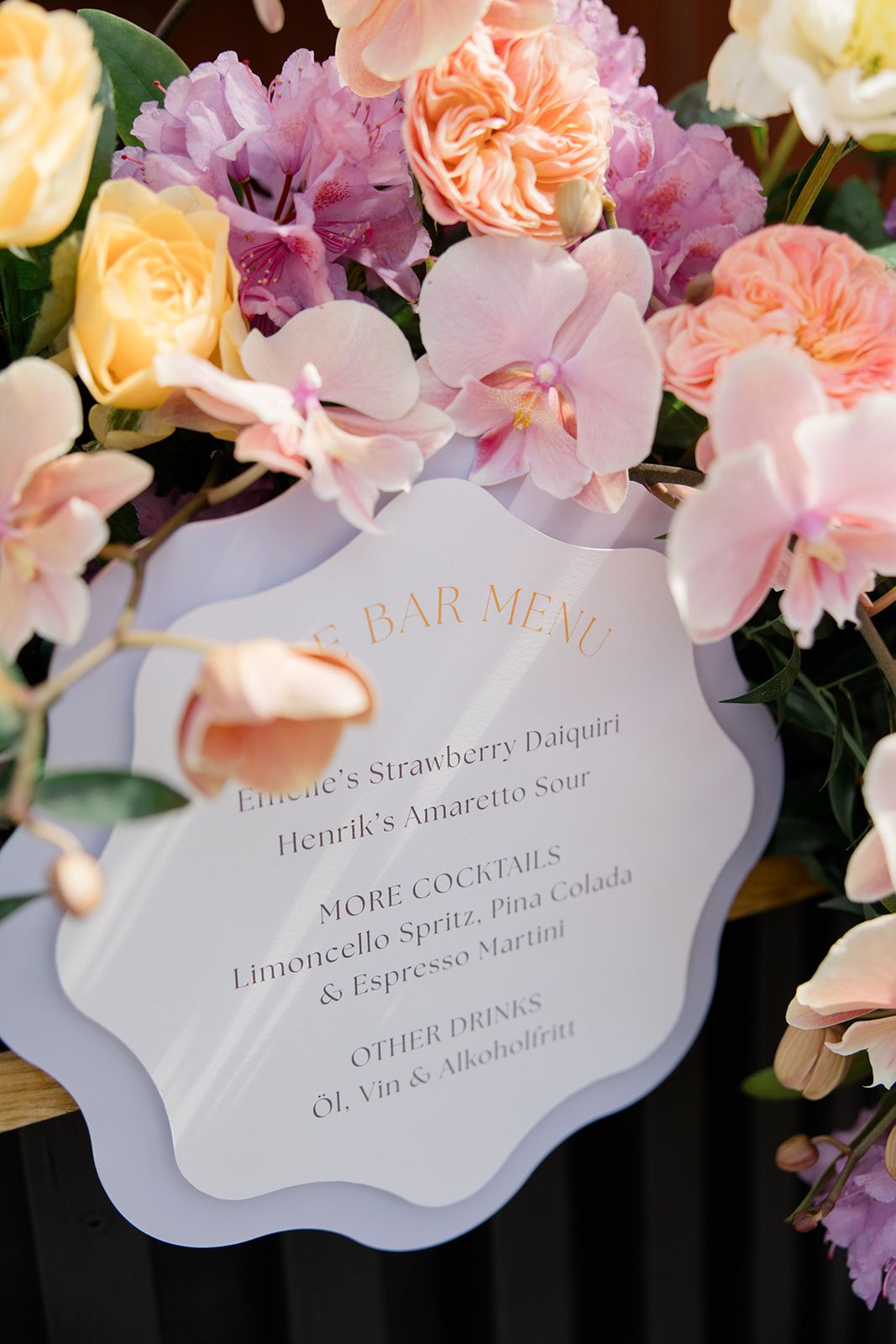 Elegant bar menu card surrounded by pink orchids, peach roses, and yellow flowers in a wedding reception setting.