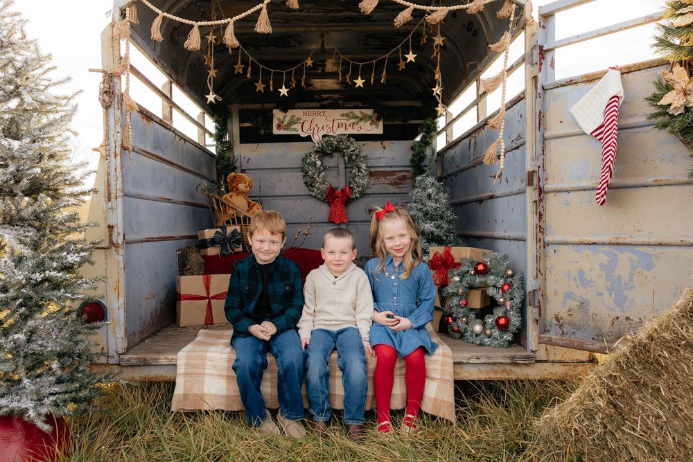 Children sit together on a rustic wooden bench in a festive holiday setting with Christmas decorations and garland.