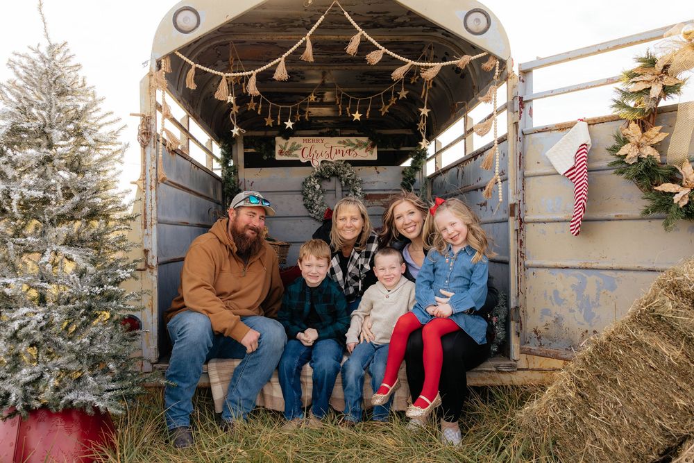 A family sits together on hay bales in a rustic barn setting decorated with holiday lights and garland.