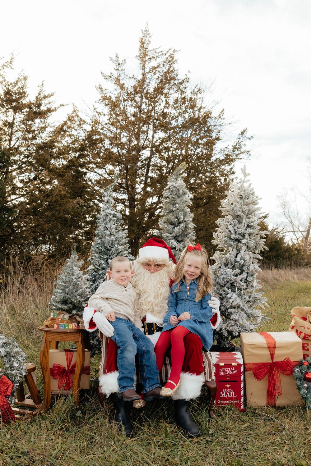 Children sitting outdoors on a winter day with flocked trees and red wrapped presents in the background.