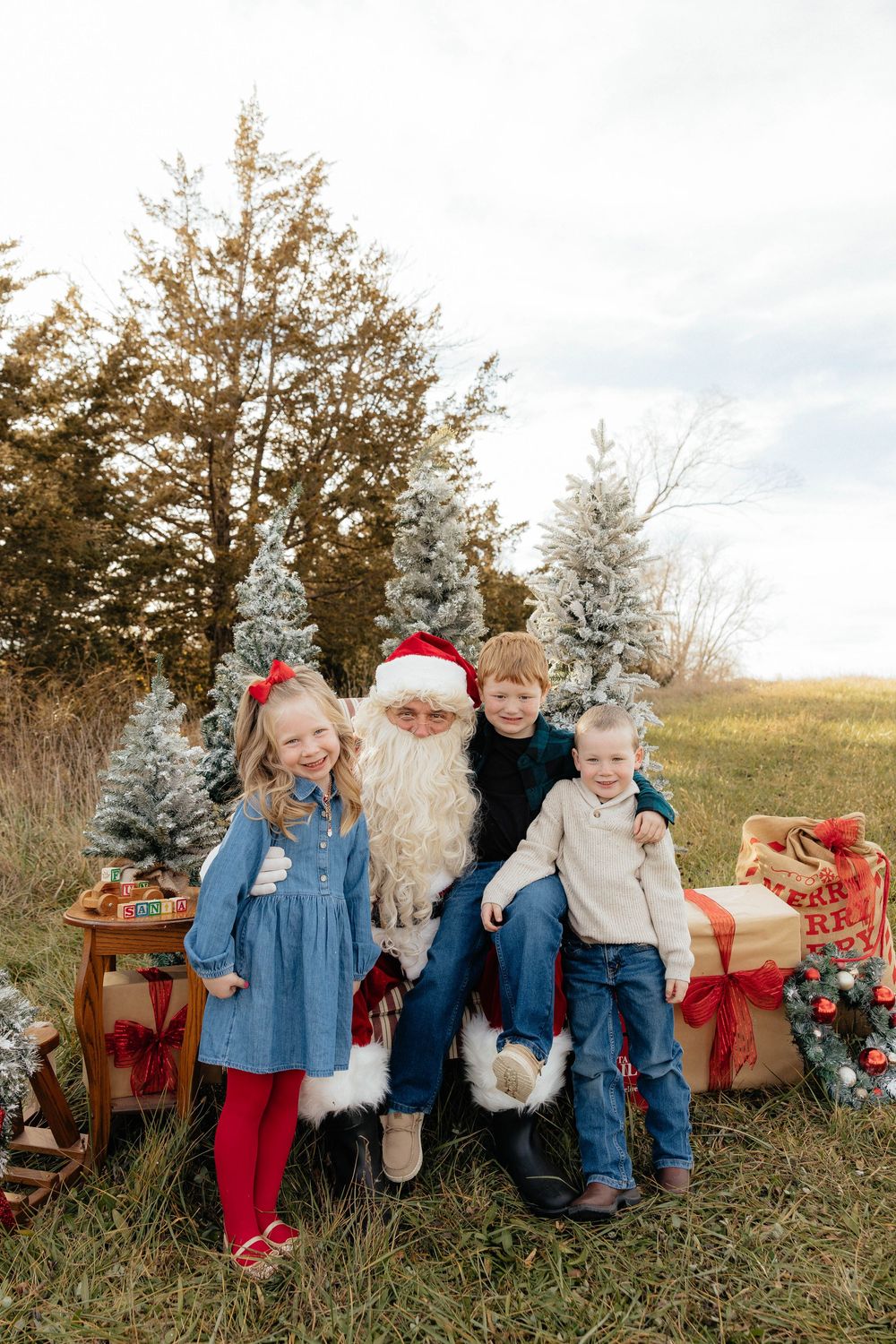 A festive outdoor Christmas scene with Santa Claus sitting on a bench with children surrounded by decorated trees and gifts.