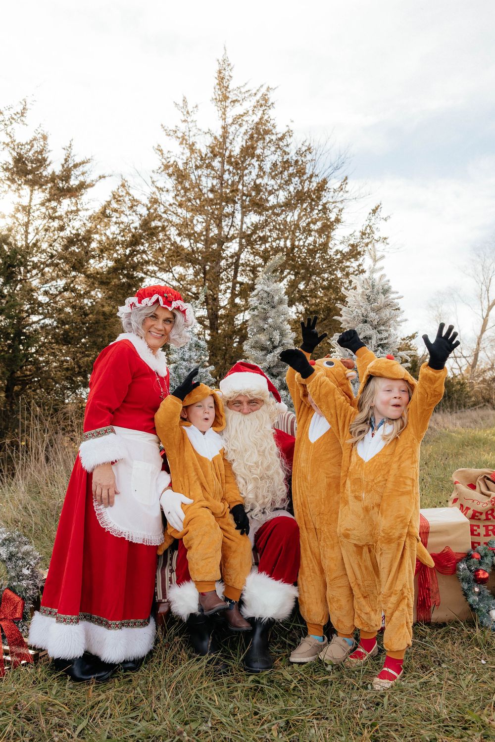 People in holiday costumes and Santa suits wave while sitting together in a grassy outdoor setting.
