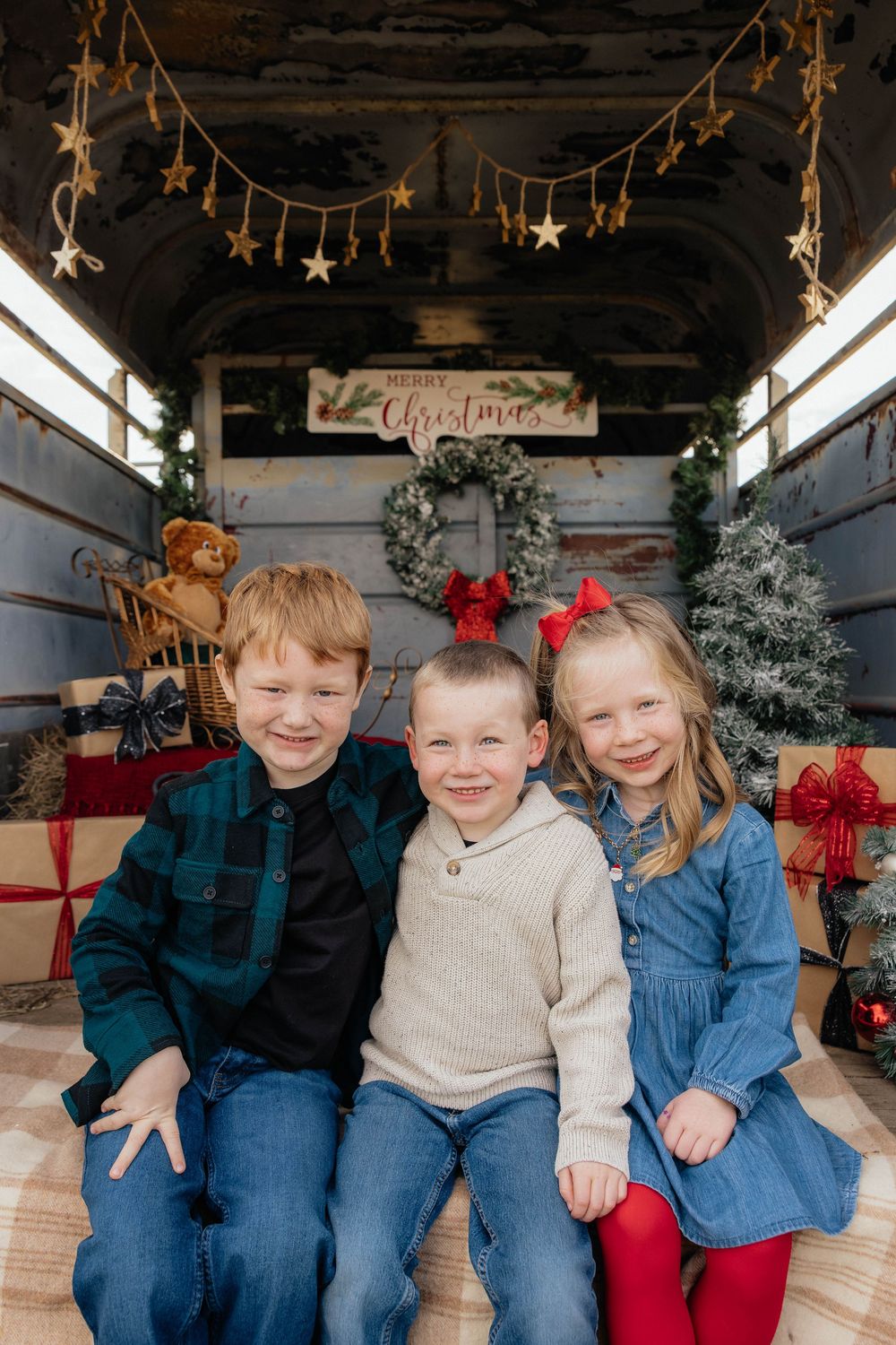Three children sitting together in a rustic holiday setting with hanging stars and festive decorations.