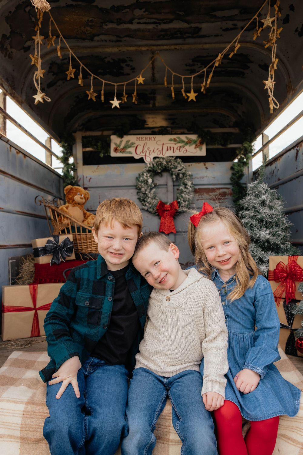 Three children sit together in a rustic wooden setting decorated with hanging stars and Christmas decor.