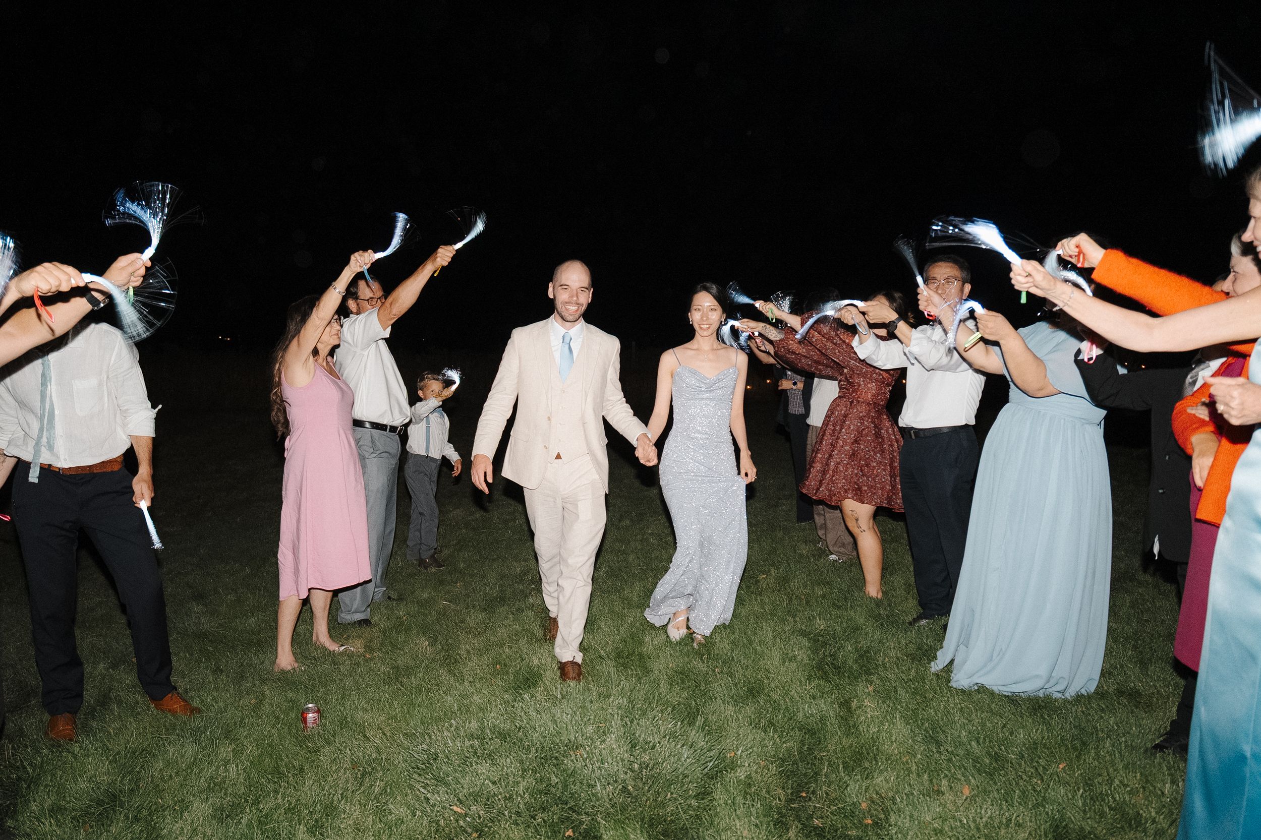 Wedding party dances in a line on grass during outdoor evening reception.
