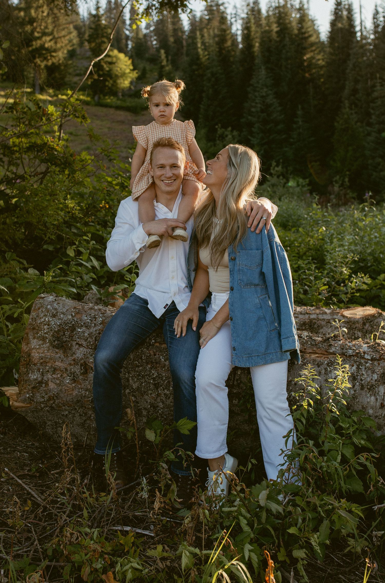 A couple shares intimate moments in a forest setting during golden hour, wearing casual white and denim outfits.