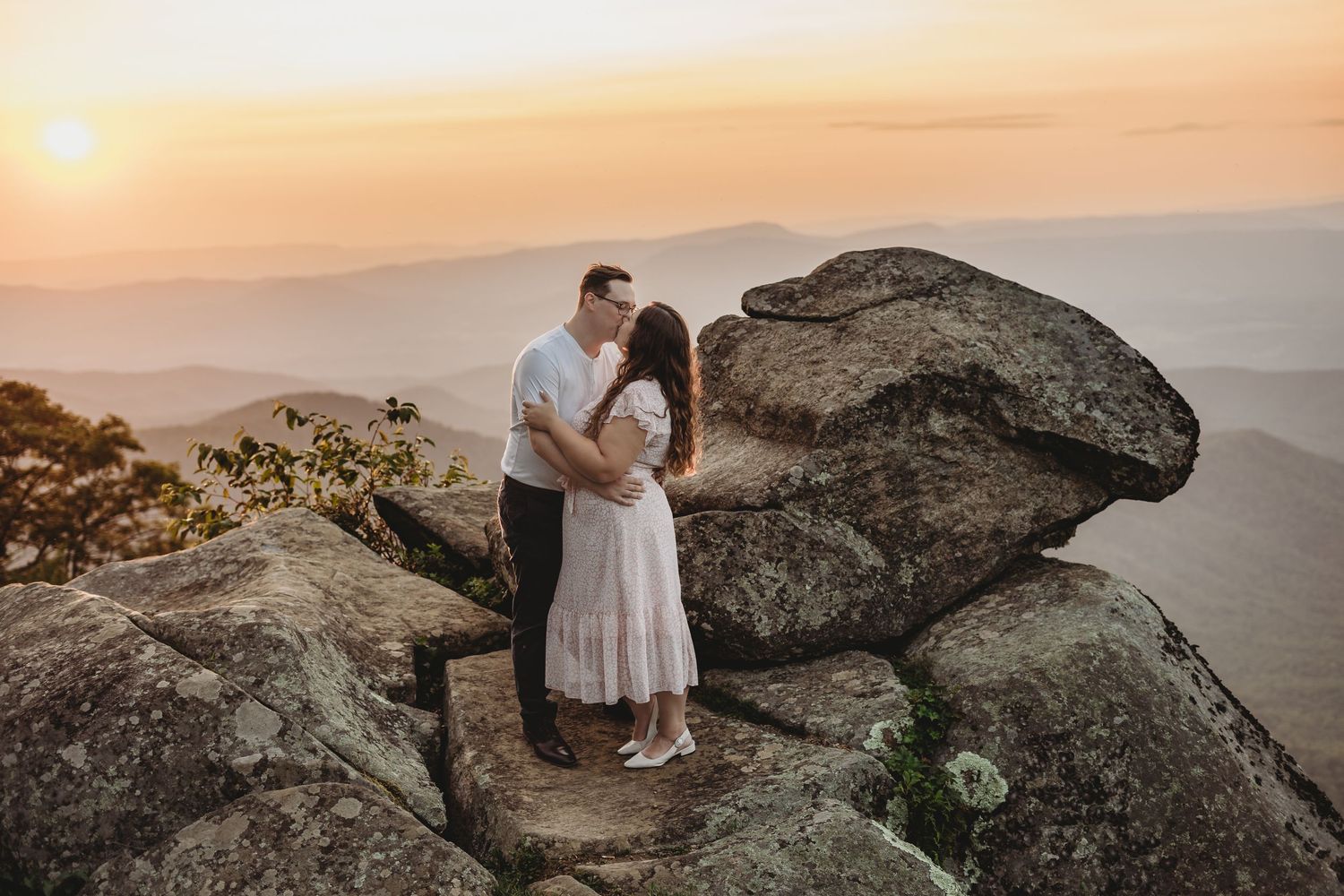 A couple celebrates their engagement on rocky mountain cliffs during a golden sunset.