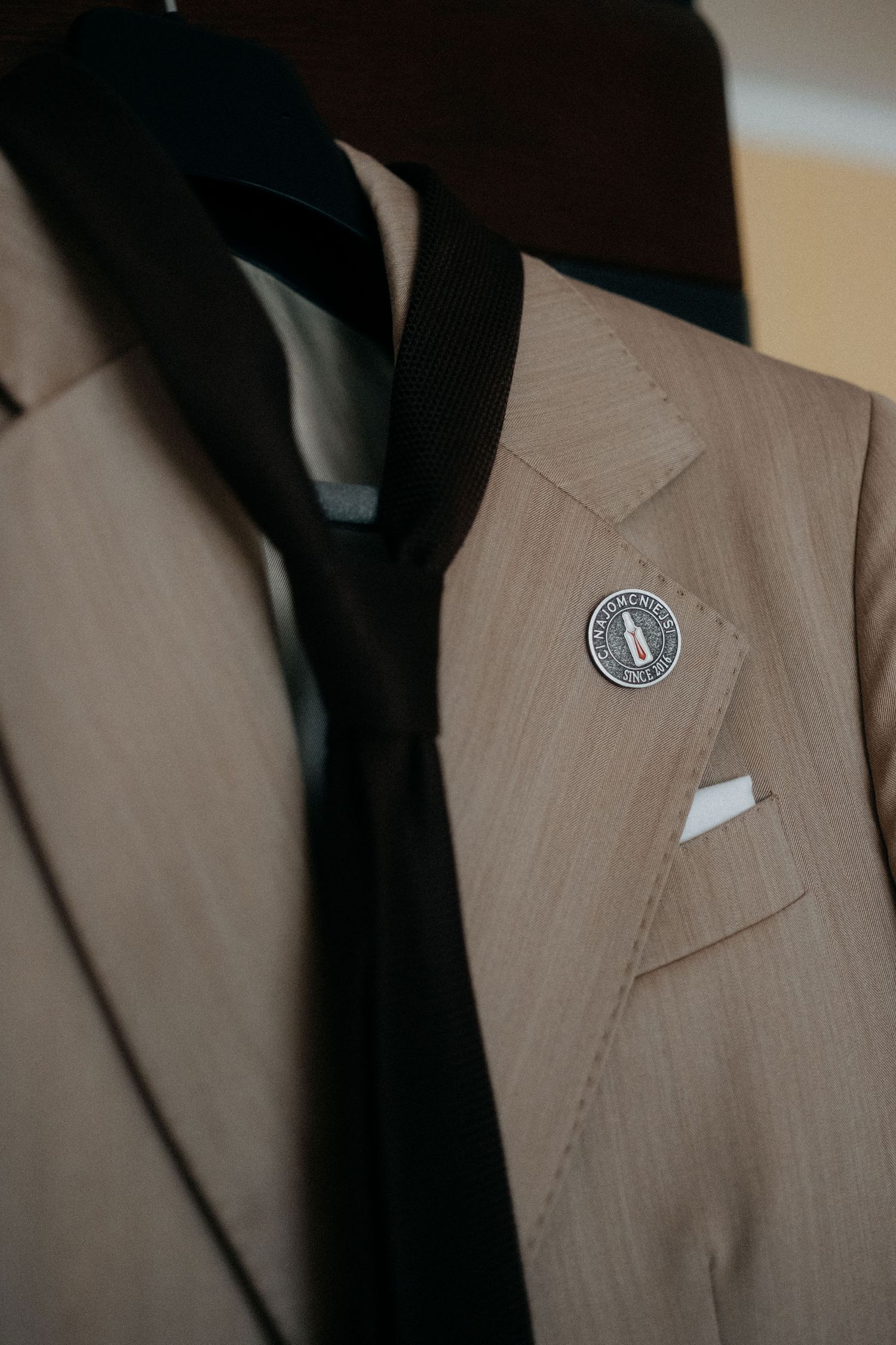Beige suit jacket with black tie and decorative lapel pin detail.