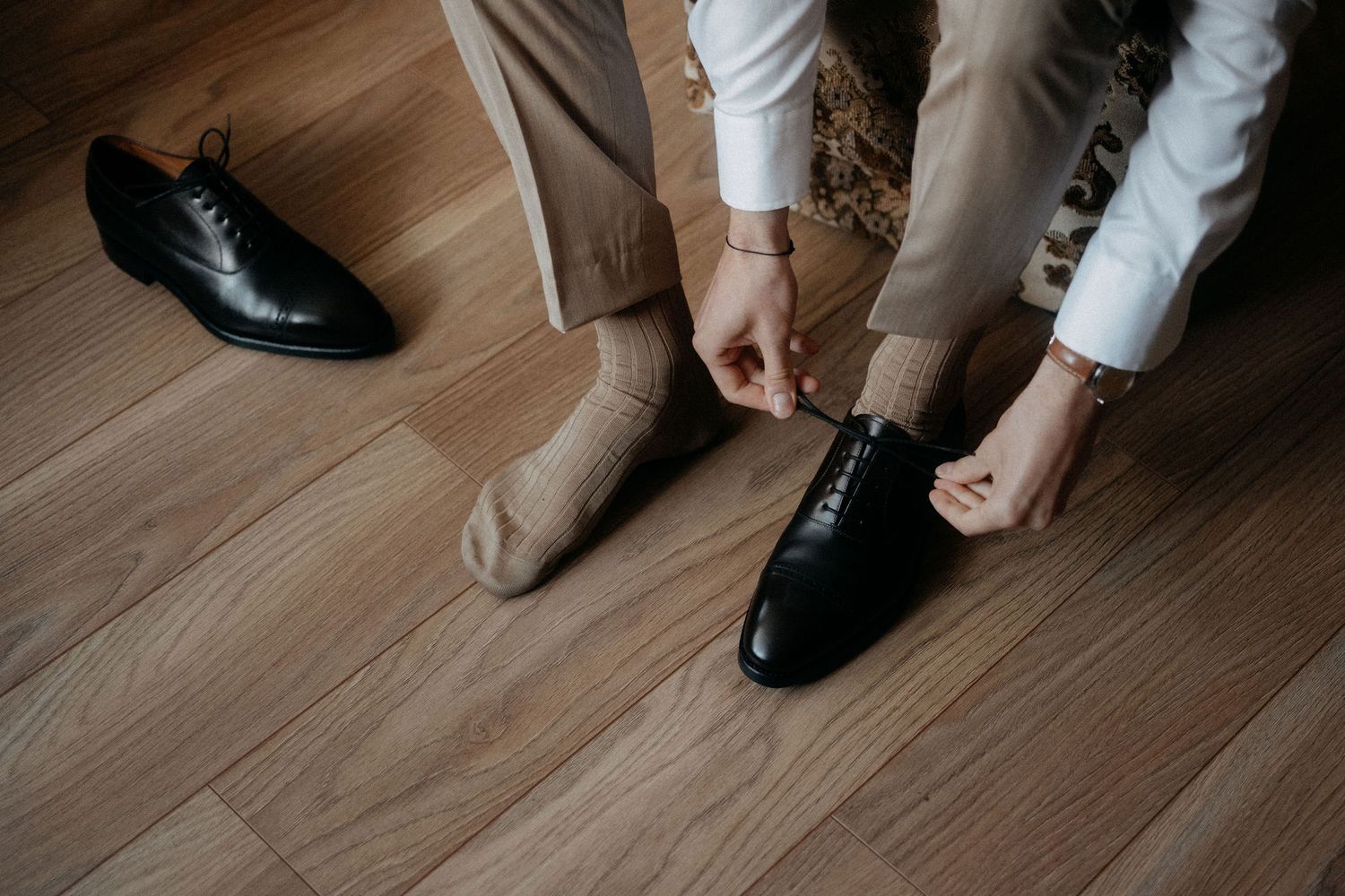 Close-up of hands tying formal black leather dress shoes on wooden floor.