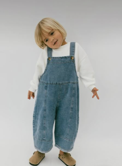 Small child in denim overalls and white sweater poses against white background.