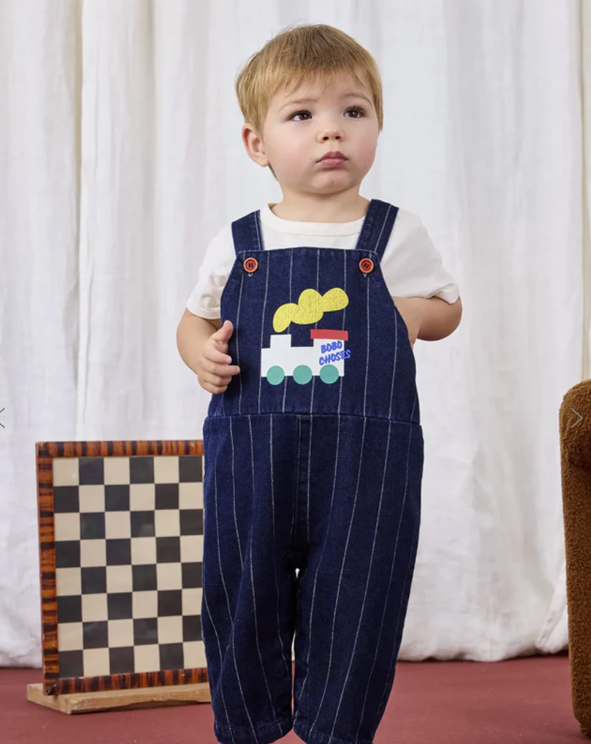 A toddler wearing navy pinstripe overalls and white shirt stands in front of a white curtain backdrop.