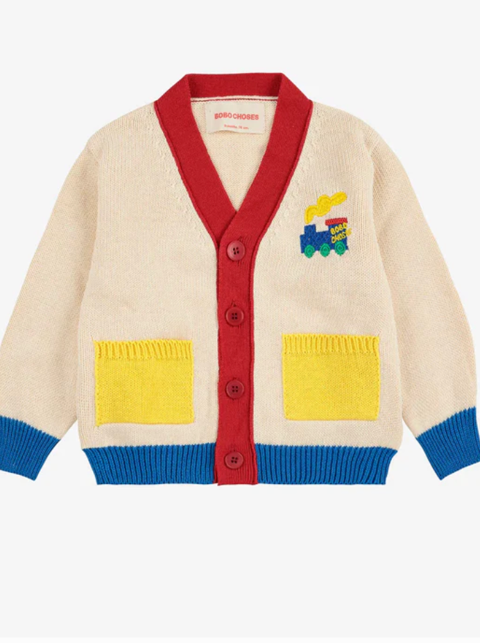 A cream colored children's cardigan sweater with red trim, yellow pockets, and a blue train applique on the chest.