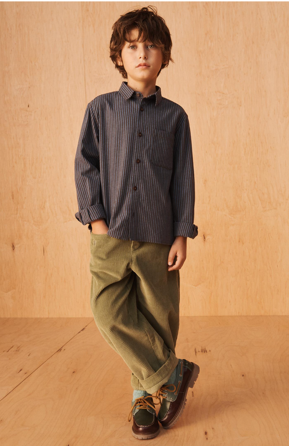 Boy in a navy striped button-up shirt, olive green corduroy pants and brown leather boat shoes standing against a light wood background.