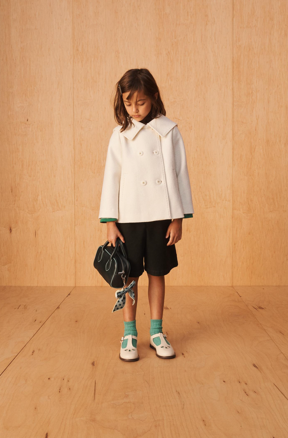 Child in a white double-breasted peacoat, black shorts, teal socks and cream T-bar shoes holding a black handbag against a plywood wall.