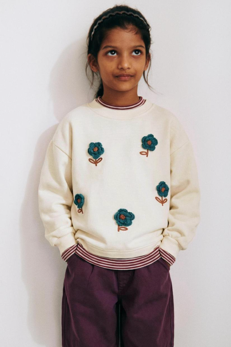 Child in a cream sweatshirt with crocheted teal flowers, striped trim and plum trousers, standing against a white wall.