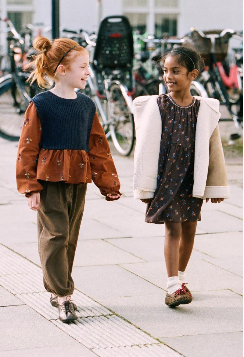 Two smiling girls in autumn outfits walk side by side on a city sidewalk with parked bicycles in the background.