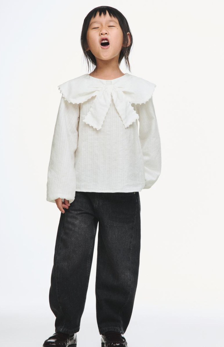 Child in a white textured blouse with a large scalloped bow collar, black wide-leg jeans and glossy black shoes, posing with eyes closed.