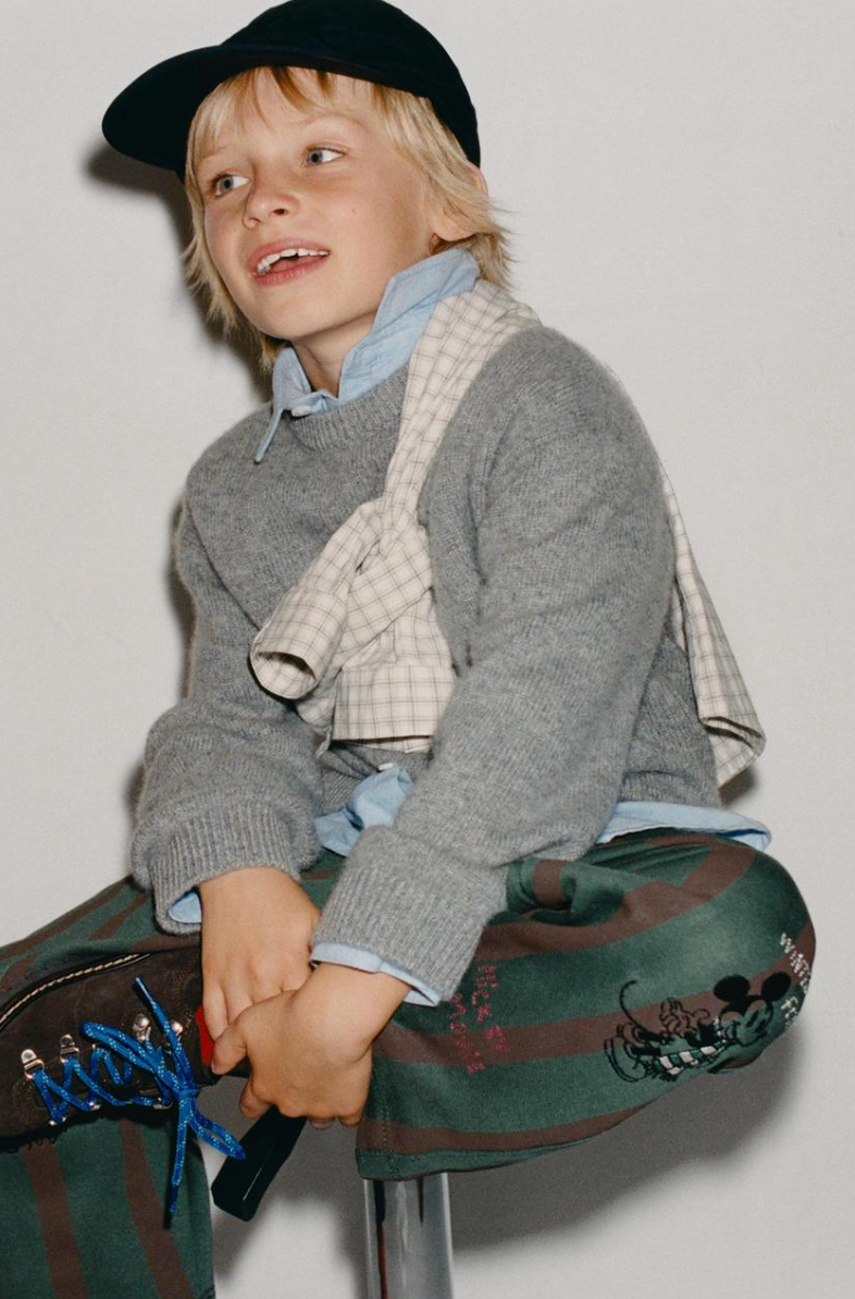 Child in a black cap, gray sweater, plaid scarf and striped green Mickey Mouse pants sits on a stool lacing blue-laced boots.