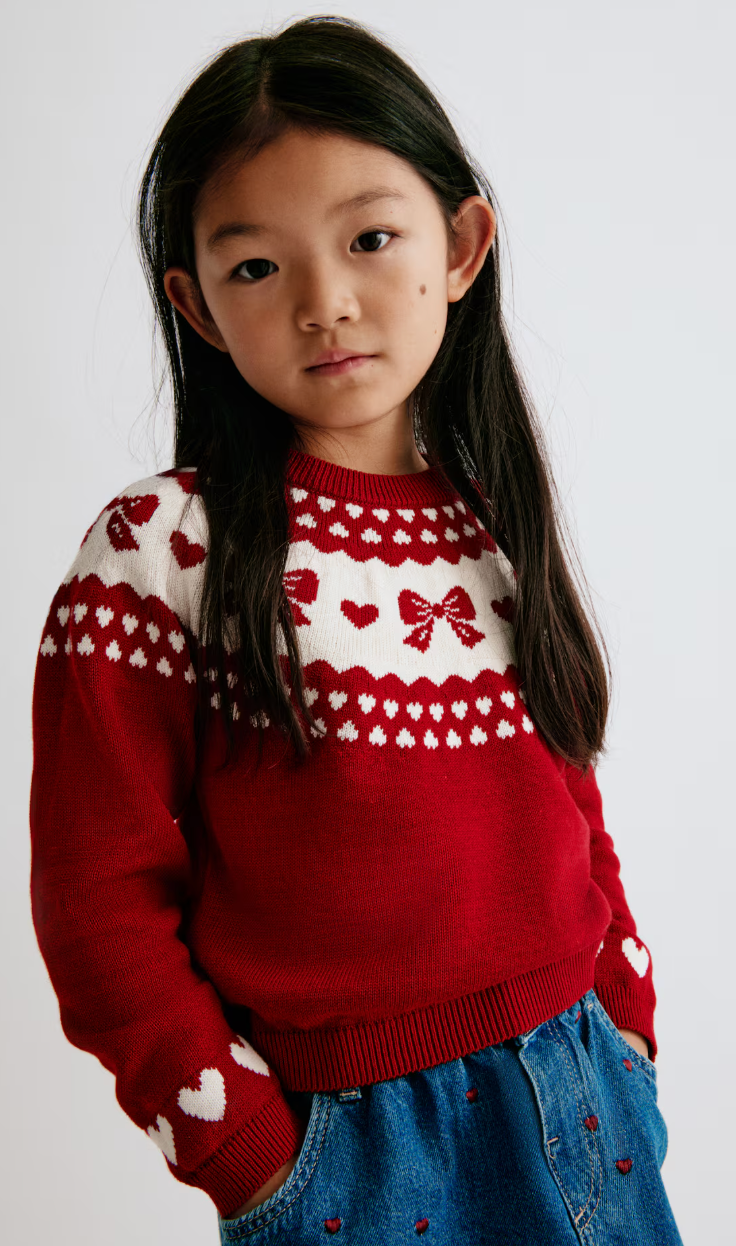 Child in a red knit sweater with white heart and bow Fair Isle yoke and heart-patch sleeves, worn with embroidered denim.