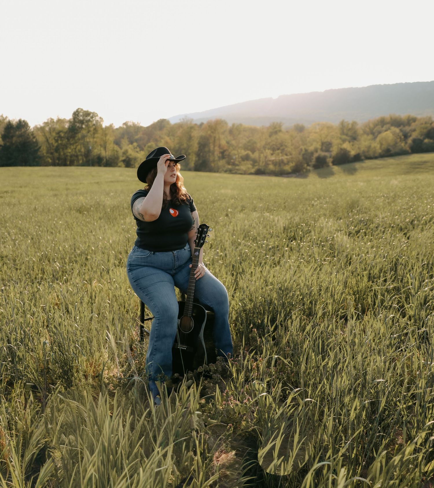 Woman in black hat and jeans holds guitar in sunlit green field with rolling hills and trees in background.