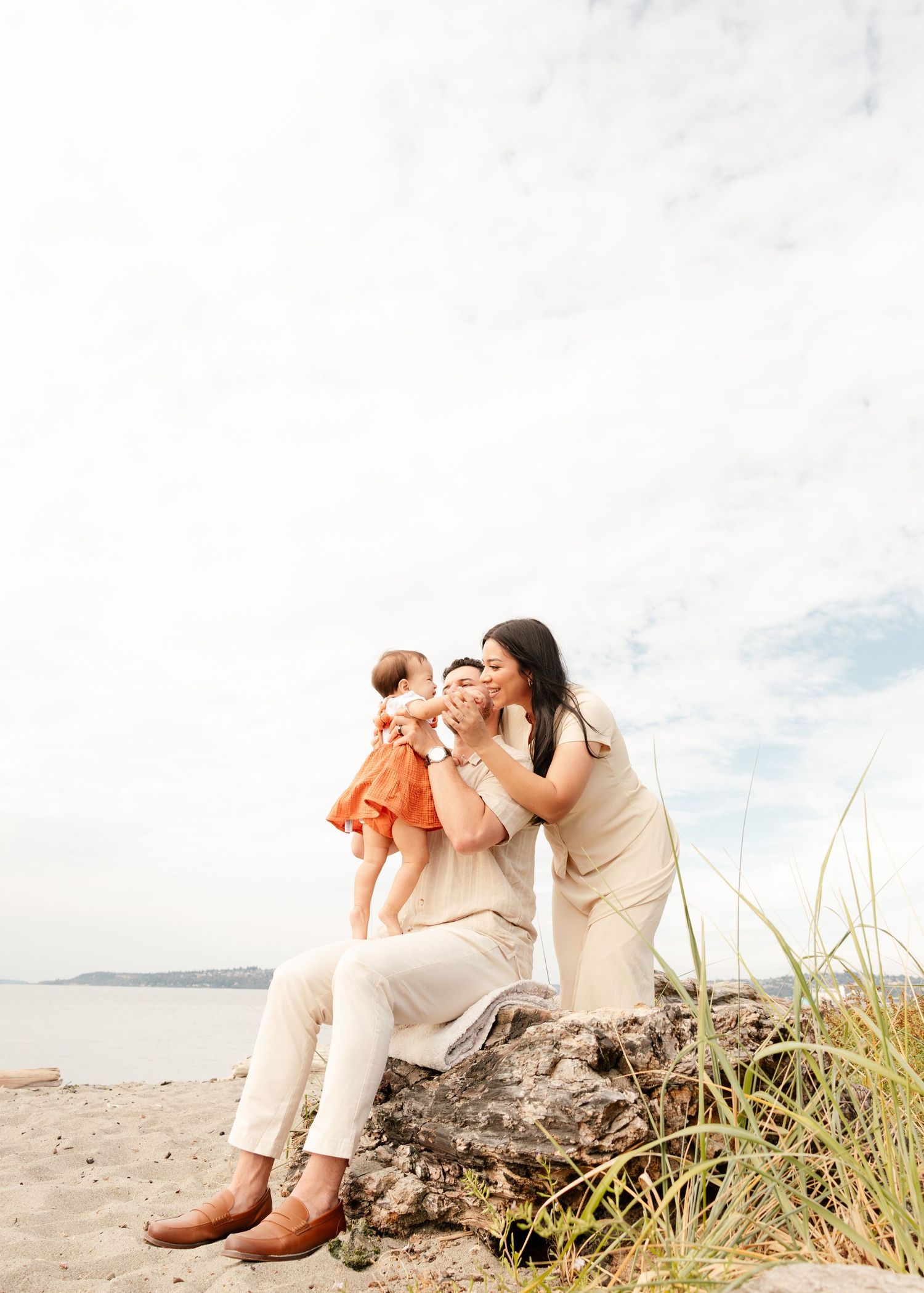 Couple in white outfits sitting together on a beach log with coastal grass and ocean views in the background.