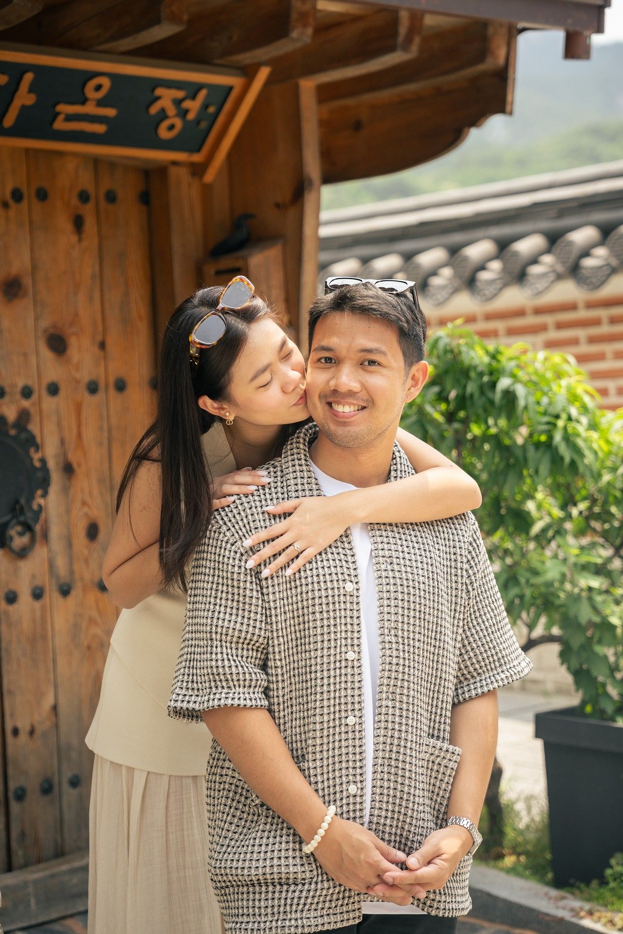 A couple embraces outside a traditional Korean building with wooden doors and brick walls on a sunny day.
