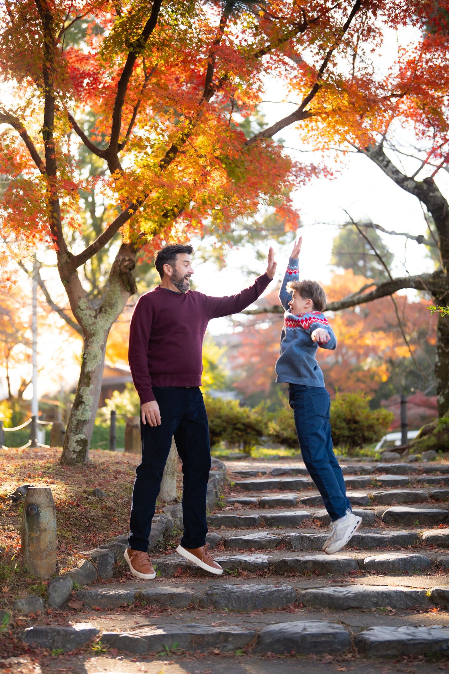 A father and son share a high-five on stone steps surrounded by vibrant autumn trees.