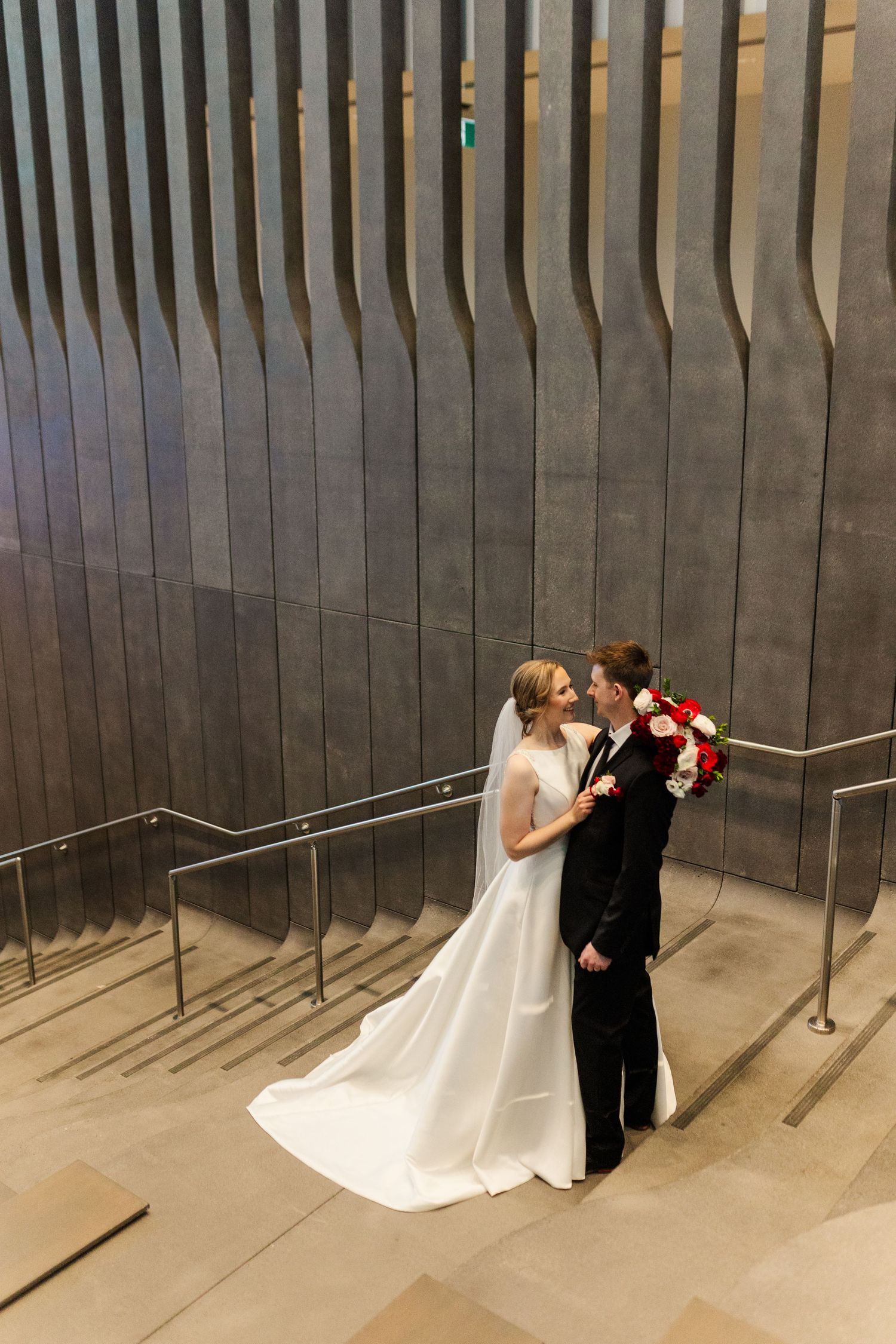 A couple in wedding attire shares a romantic kiss in front of a modern architectural wall with vertical metal slats.