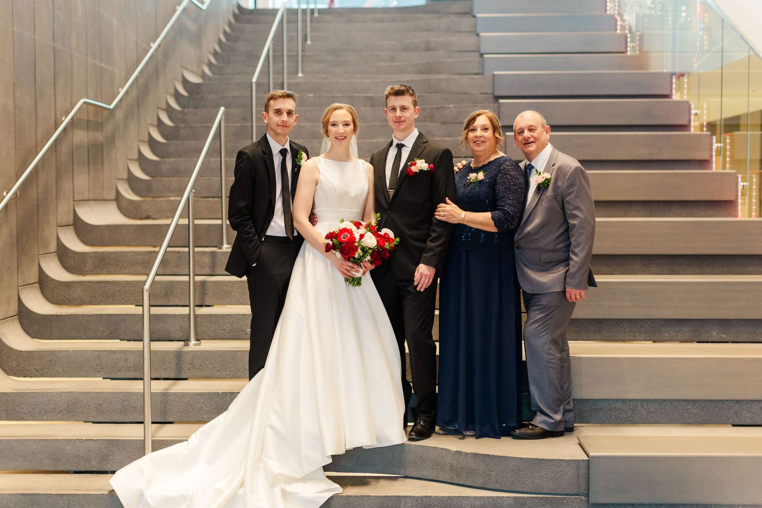 A formal wedding party poses on modern stairs with the bride in white holding a red bouquet.