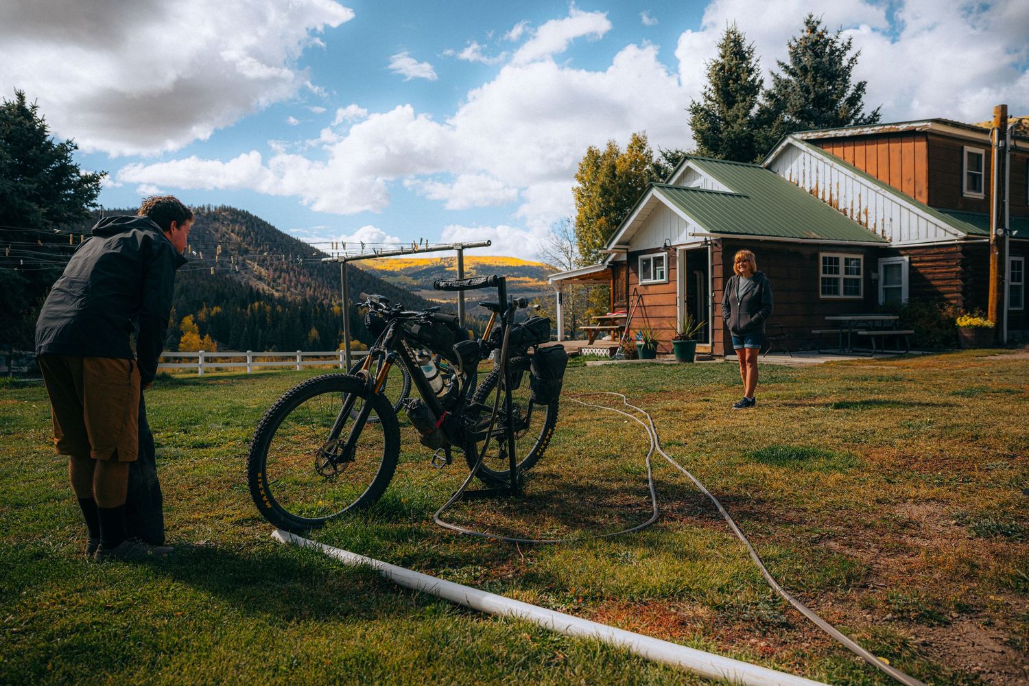 A person rides a bicycle through a rural landscape with rustic cabins and mountain scenery in the background.
