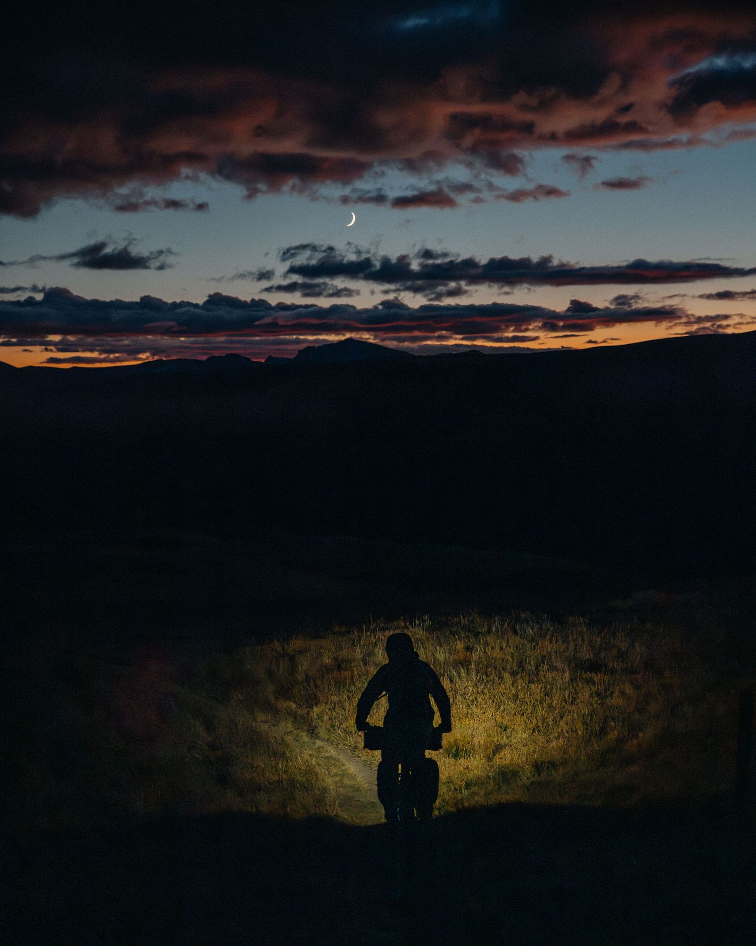 A silhouetted figure illuminated by flashlight stands in a grassy field under a dramatic sunset sky.