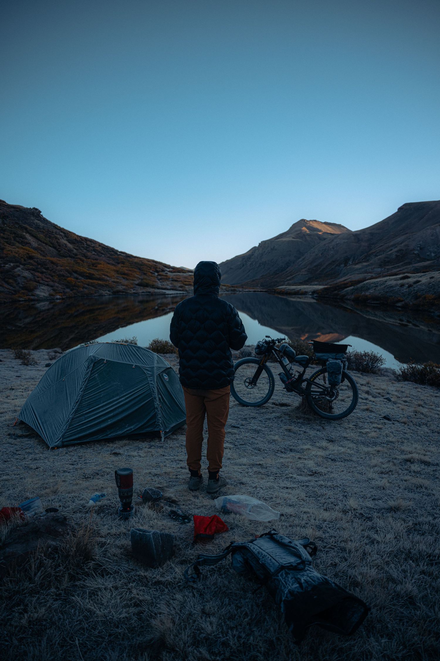A campsite with tent and bicycle overlooks a serene mountain lake at dusk.